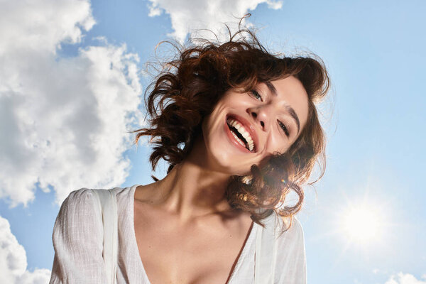 A young woman with wavy brown hair smiles joyfully under a clear blue sky on a summer day.