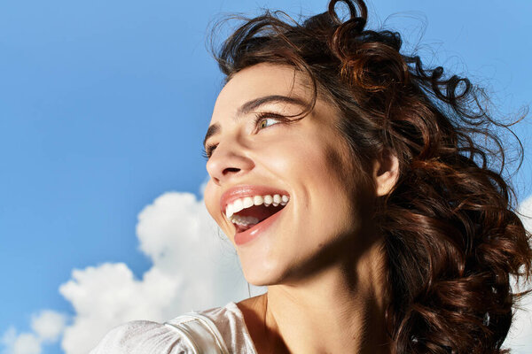 A young woman with wavy brown hair smiles brightly against a clear blue sky on a sunny day.
