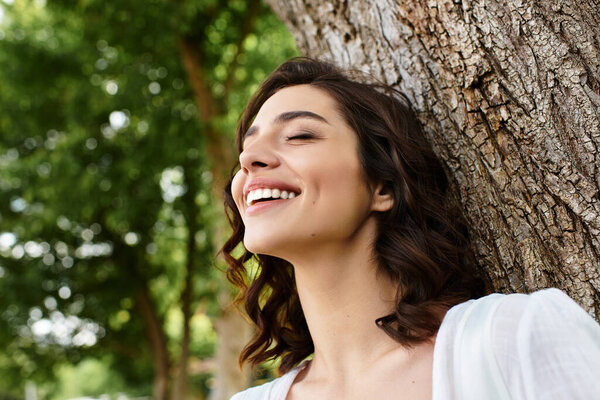 Wavy brown-haired woman smiles happily while resting against a tree in the park.
