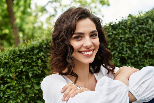 Молодая женщина с волнистыми каштановыми волосами тепло улыбается, обнимая лето на открытом воздухе..