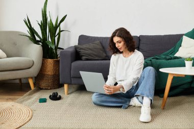 Evde rahat bir şekilde otururken laptopuna odaklanmış kıvırcık saçlı genç bir kadın..