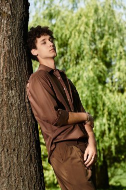Şık bir genç adam güneşli park ortamının keyfini çıkarırken bir ağaca yaslanır..