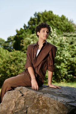 Şık yaz kıyafetleri içindeki genç adam yemyeşil bir kayanın ortasında, doğanın tadını çıkartıyor..