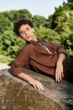 Kıvırcık saçlı genç adam yazın yemyeşil bir kayanın üzerinde rahatlar..