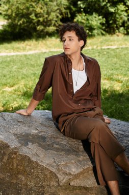 Şık bir kıyafet giyen genç bir adam, parktaki bir kayanın üzerinde dinleniyor ve yaz güneşini emiyor..