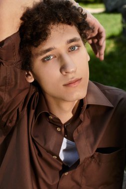 Kıvırcık saçlı genç bir adam yemyeşil bir parkta gevşiyor, yaz modası ve tarzı somutlaştırıyor..