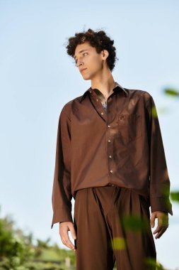 Kıvırcık saçlı, şık bir yaz kıyafeti giymiş genç bir adam yeşil bir parkta dışarıda dinleniyor..