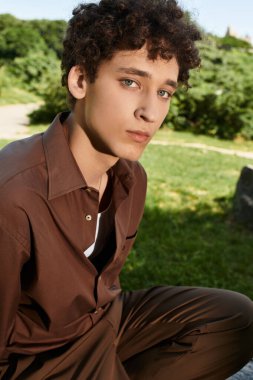 Kıvırcık saçlı genç bir adam, son moda yaz kıyafetleri içinde parkta rahatlıyor..