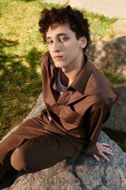 Kıvırcık saçlı genç bir adam canlı bir parkta bir kayanın üzerinde gevşiyor, yaz modasını kucaklıyor..