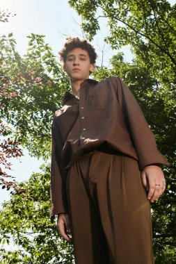 Kıvırcık saçlı genç bir adam, bereketli bir parkta dinlenirken yaz modasına sarılır..