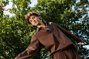 Kıvırcık saçlı genç adam yemyeşil ortamda şık bir yaz kıyafetiyle dışarıda dinleniyor..