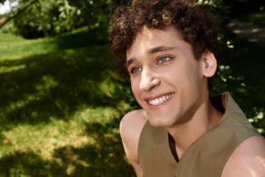 Kıvırcık saçlı genç bir adam açık havada güneşli yeşil bir parkta dinlenirken gülümsüyor..