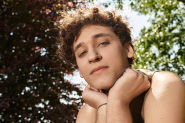 Kıvırcık saçlı genç bir adam bereketli bir yaz parkı atmosferinde rahatlar ve neşe ifade eder..