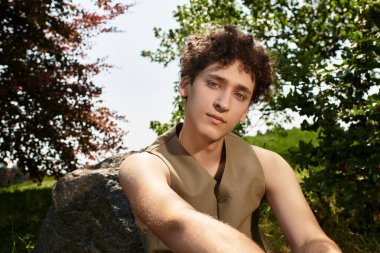 Kıvırcık saçlı genç adam dışarıda rahatlıyor ve şık yaz kıyafetini parkta sergiliyor..