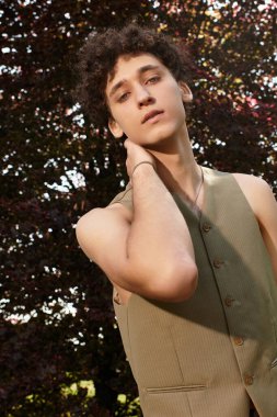 Kıvırcık saçlı genç bir adam yeşil bir parkta dinleniyor, son moda yaz kıyafetleriyle güneşin tadını çıkarıyor..