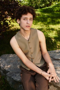 Kıvırcık saçlı genç bir adam, parkta şık bir yaz kıyafeti giymiş bir taşın üzerinde dinleniyor..