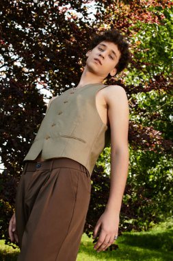 Kıvırcık saçlı genç adam açık havada rahatlıyor, bereketli yeşillikler arasında moda olan yaz modasını sergiliyor..