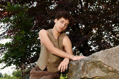 Kıvırcık saçlı genç bir adam yeşil bir parkta rahat ve şık bir yaz kıyafeti giyiyor..