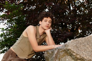 Tarz sahibi genç bir adam yeşil bir parkta, eşsiz yaz kıyafetleri ve bukleleriyle hava atıyor..