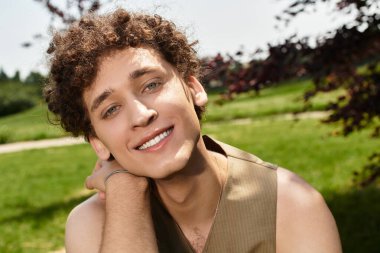 Kıvırcık saçlı genç bir adam parkta rahatlıyor, son moda yaz kıyafetlerini sergiliyor ve gülümsüyor..