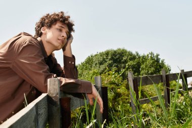 Kıvırcık saçlı genç adam güneşli bir günde, yemyeşil bir parkta, ahşap bir çitin yanında rahatlar..