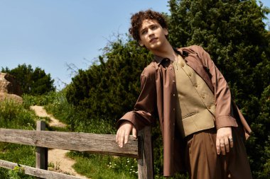 Kıvırcık saçlı genç adam, etrafındaki doğanın güzelliğinin tadını çıkarırken ahşap bir çitte dinleniyor..