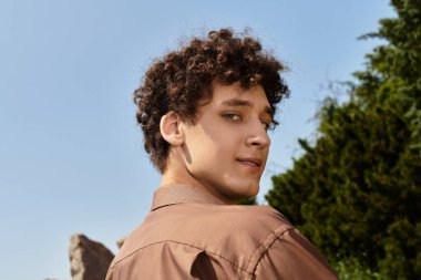 Kıvırcık saçlı genç adam yeşil bir parkta güneşli yaz kıyafetlerini sergiliyor..