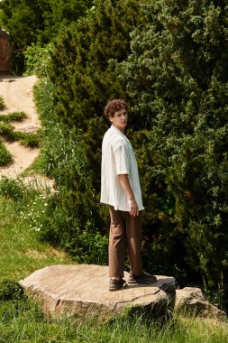 Kıvırcık saçlı genç bir adam şık bir yaz elbisesi içinde canlı bir parkı keşfediyor..