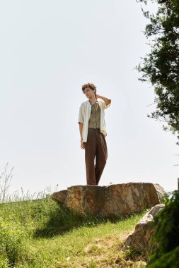 Kıvırcık saçlı genç bir adam güneşli bir günü kucaklayarak canlı bir parkta bir kayanın üzerinde dinleniyor..