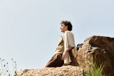 Kıvırcık saçlı şık bir genç adam yemyeşil bir parkta bir kayaya yaslanır ve gün ışığını hisseder..