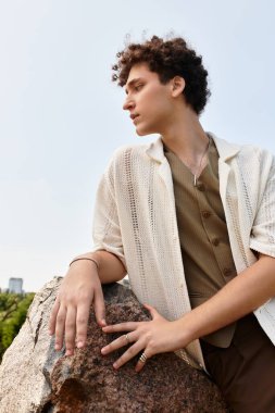 Kıvırcık saçlı, şık yaz kıyafetleri giymiş genç bir adam yemyeşil bir parkta rahatlıyor..