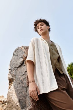 Kıvırcık saçlı genç adam yeşil bir parkta dinleniyor, açık bir gökyüzü altında yaz modasını sergiliyor..