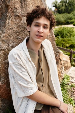 Kıvırcık saçlı genç adam parkta yemyeşil bir kayanın üzerinde rahatlıyor..