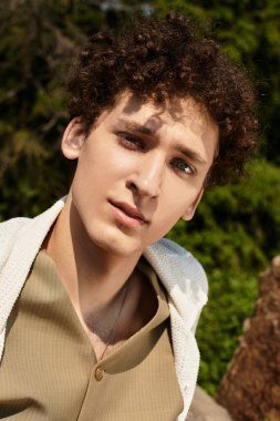 Kıvırcık saçlı genç bir adam şık yaz kıyafetiyle parkta güneşli bir günün tadını çıkarıyor..