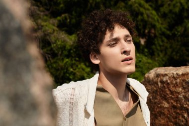 Kıvırcık saçlı genç bir adam hareketli bir parkta gevşeyip son moda yaz görüntüsünü sergiliyor..