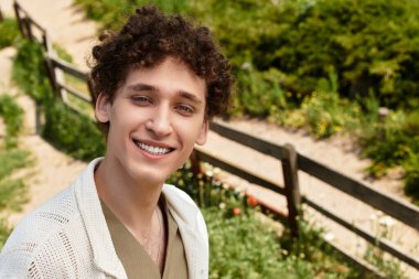 Kıvırcık saçlı genç bir adam bir parkta rahat, modaya uygun bir yaz kıyafeti giyiyor ve gülümsüyor..