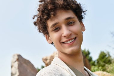 Kıvırcık saçlı genç bir adam yeşil bir parkta yaz havasını kucaklıyor, açık havanın tadını çıkarıyor..