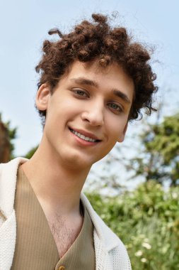 Kıvırcık saçlı genç bir adam yemyeşil bir parkta rahat, şık bir yaz kıyafeti giymiş..