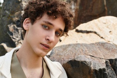 Kıvırcık saçlı genç bir adam lüks bir park ortamında dinlenirken şık bir yaz kıyafeti sergiliyor..