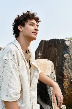 Kıvırcık saçlı, şık bir yaz kıyafeti giymiş genç bir adam enerjik bir parkta dinleniyor ve açık havanın tadını çıkarıyor..