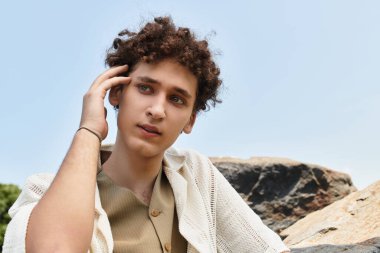 Kıvırcık saçlı genç adam yeşil bir parkta yaz atmosferini kucaklıyor, modern moda sergiliyor..