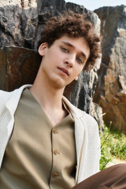 Kıvırcık saçlı genç bir adam lüks bir parkta, şık bir yaz kıyafeti içinde rahatlar..