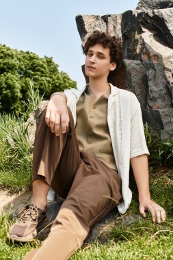 Kıvırcık saçlı genç bir adam güneşli park atmosferinde, bir kayanın üzerinde dinleniyor..