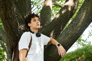 Beyaz tişörtlü genç bir adam bir ağaca yaslanır, parkta yaz esintisinde ıslanır..