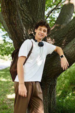 Beyaz tişörtlü kıvırcık saçlı genç bir adam, parlak bir yaz parkında bir ağacın altında dinleniyor..