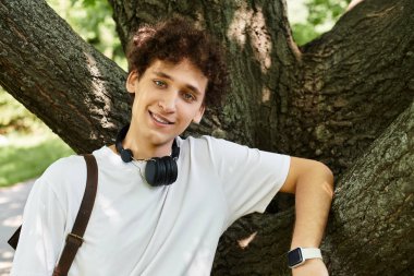 Kıvırcık saçlı genç bir adam sıcak bir yaz gününde bir ağacın altında beyaz tişört giyerek rahatlıyor..