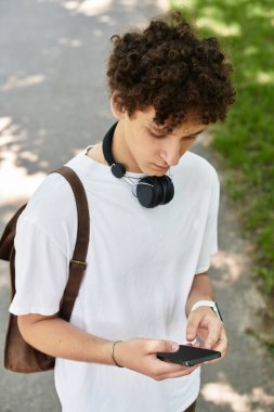 Yeşil bir parkta, kıvırcık saçlı genç bir adam telefonunu kontrol ediyor, yaz havasını içine çekiyor..