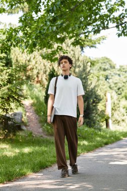 Kıvırcık saçlı genç bir adam canlı yeşil bir parkta beyaz bir tişört giyerek dolaşıyor..