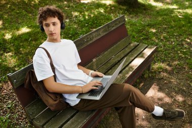 Kıvırcık saçlı genç bir adam dışarıda vakit geçirmekten hoşlanıyor, bir bankta otururken dizüstü bilgisayarla çalışıyor..