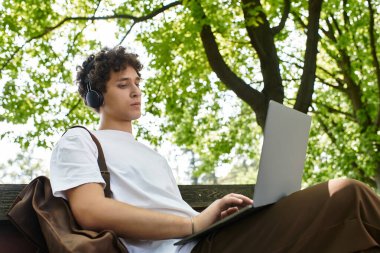 Kıvırcık saçlı genç bir adam yeşil bir parkta rahatlıyor, doğanın ortasında laptopuna odaklanıyor..